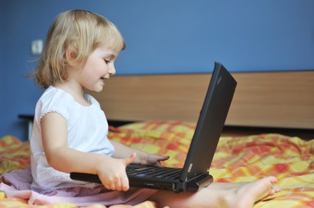 cheerful little girl with laptop sits on bed of parentsの写真素材