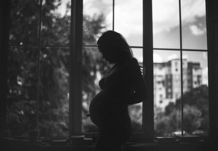 The future mother looks at her belly waiting for the special day near the window and is photographed in contre-jourの写真素材