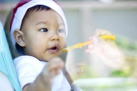 a baby asian in a Santa Claus hat and looking at camera is sitting in a chair and eating  Merry...