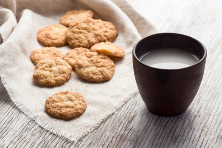 Oatmeal cookies on a linen a napkin with milk in ceramic cup on a light wooden background. Breakfast in the village.の写真素材