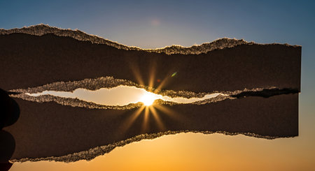 Sunburst visible through torn paper held against sunset skyの素材