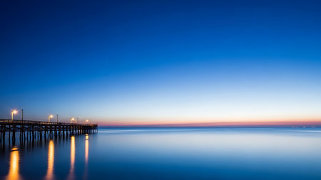 Pier with illuminated lights reflecting on calm ocean water at dawn, vast blue skyの素材