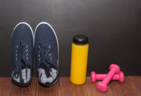 Blue fitness shoes, orange juice and pink weights on wooden floor in front of black deskの写真素材