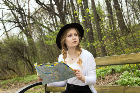 Blonde woman in black hat holding map, sitting on bench in the forest. Tourist planning her route in park.の写真素材