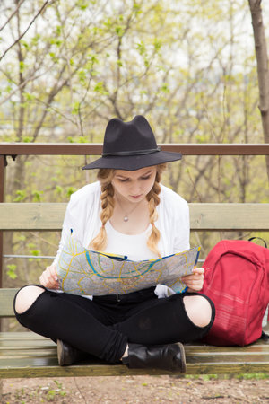 Attractive blonde woman in black jeans, white coat and blackhat looking at map, while siting on bench in the park. Red backpack stands near.の写真素材