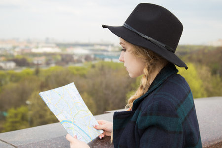 Cute blonde woman in black hat looking at sightseengs map, while standing at observation deck.の写真素材