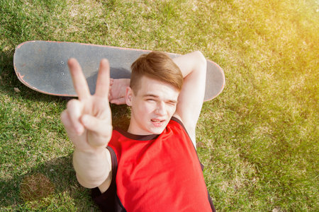 Young man lying on grass in park shows peace sign, skateboard near, summertimeの写真素材