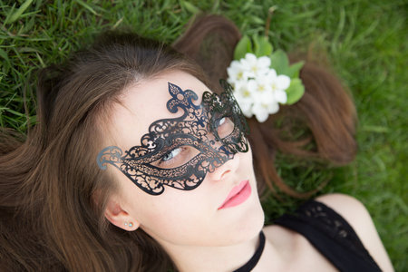 close up portrait of sensual beautiful brunette young lady  wearing mask  lying on green grass with flower in her hairの写真素材