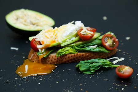 Healthy toast with avocado, tomatoes, spinach and poached egg. Dark background, half of cheese avocado behind.の写真素材