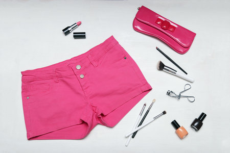Top view of female fashion accessories for woman. Pink shorts, clutch, lipstick, nail polish on white wooden background.の写真素材