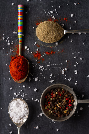 Old spoons with peppercorn, ground pepper, salt and red paprika powder on black background. Flat lay. Top view. Food concept. Dark mood food photography.の写真素材