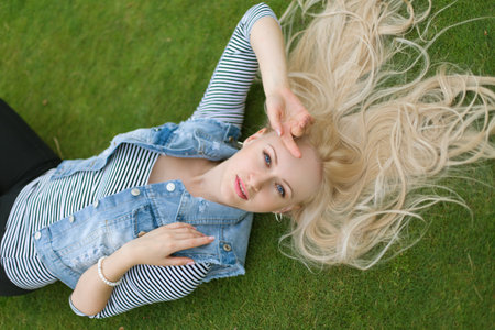 Blonde woman in jeans vest is lying on the green grass with untressed hairの写真素材