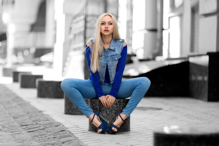 Beautiful blonde woman in jeans costume and high heels sitting outdoor on the stone bench - photo with desaturated backgroundの写真素材