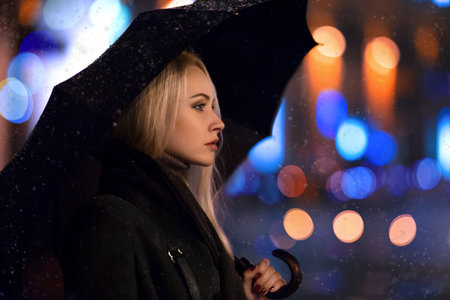 Beautiful girl with umbrella posing on the winter street in the lights of the evening city with snow and bokeh. Portrait with loneliness sad emotions.の写真素材