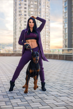 Beautiful fitness brunette girl with perfect legs posing on the city square in sexy sports costume with huge Doberman dog in the rays of setting sun.の写真素材