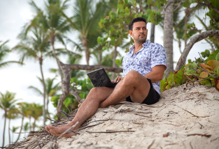 Successful freelancer businessman working with laptop on the tropical beach in the shadows of palm trees.の写真素材