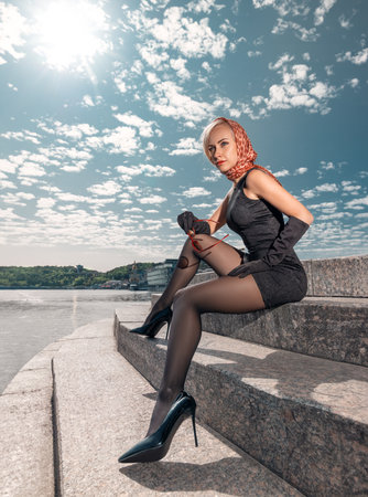 Beautiful blonde girl in black dress with perfect legs in pantyhose and shoes with high heels posing outdoors near the river bridge in the rays of sun. Street fashion photo. Wide angle shot.の写真素材