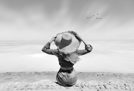 A woman with a big hat with a desert sea view watches flying crane birds coming home. Black and white conceptual minimalism shot.の写真素材