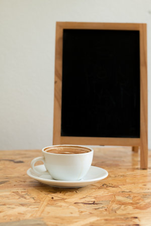 A white cup of hot coffee on wooden table,A refreshment before work time or refreshment in relaxing time in a break with empty black board for write menu in coffee shop.の写真素材