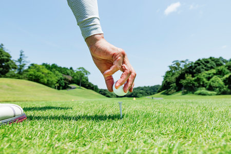 Hands placing a golf ball on a tee on a teeing ground with fresh greeneryの写真素材