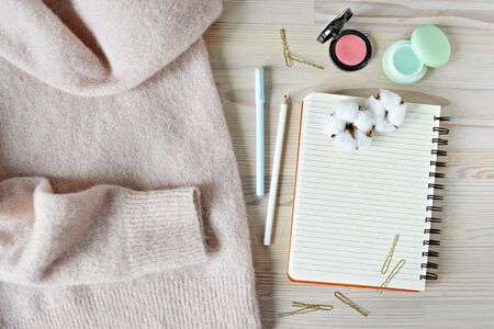 Light pink wool sweater, black spiral notebook, mint pen, white pencil, golden hairpins, pink blush, lip balm and dried cotton flowers on the wooden deskの写真素材