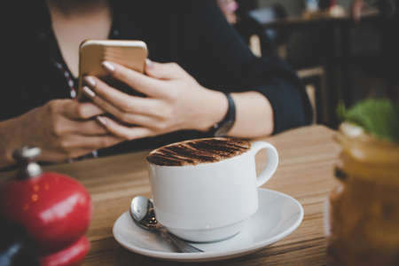 Woman typing text message on smart phone in a coffee shop. Cropped image of young woman sitting at a table with a coffee using mobile phone.の写真素材