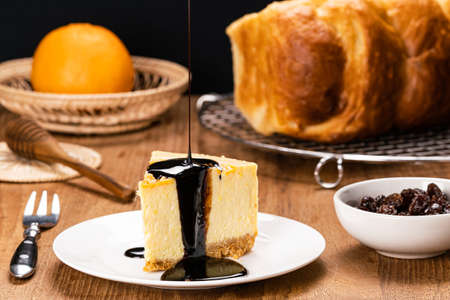 Pouring chocolate on delicious homemade mango cheesecake in white ceramic dish with metal fork, honey dipper, loaf of bread on cooling rack, ripe orange in bamboo tray and raisin in white ceramic cup.の写真素材