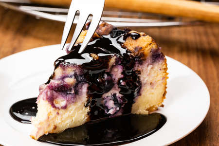 Using metal fork taking a bite from a piece of homemade blueberry and crumble cheesecake topping with chocolate in white ceramic plate on wooden table.の写真素材