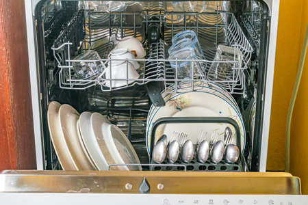 Front view of opened dish washer with clean dishes, glassware and other kitchen utensils after...