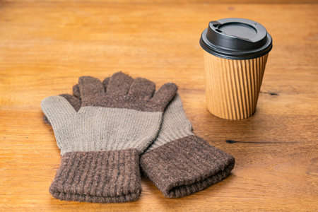 High angle view a pair of cloth gloves and a black plastic cap paper takeaway cup of hot coffee on wooden table in winter season.の写真素材
