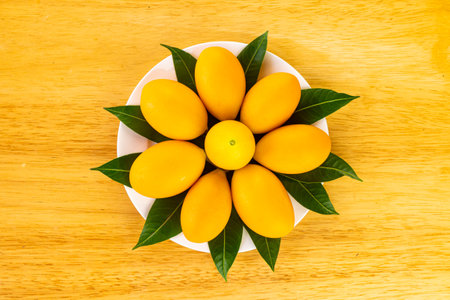 Top view of ripe sweet yellow marian plum or Bouea macrophylla griffith or maprang or mayongchid with green leaves decorated in white ceramic dish on wooden table.の写真素材