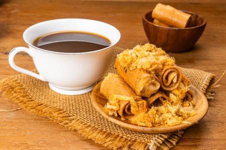 High angle view pile of sweet crispy coconut roll with dried shrodded pork in wooden plate with a cup of hot black coffee on sack cloth on wooden table.の写真素材