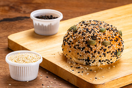 High angle view of homemade burger bun sprinkled with white and black sesame and pumpkin seeds on wooden board with cups of sesame on wooden background.の写真素材