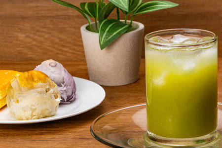 Closeup view of cold green guava juice with ice cube in transparent glass  on glass saucer with multi colored chinese pastry moon cake and layered cake in white ceramic dish on wooden table.の写真素材