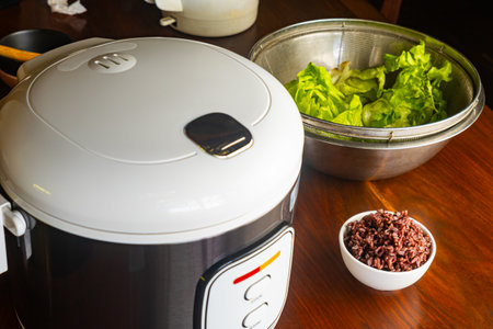 High angle view of electric rice cooker, steamed brown riceberry in white ceramic bowl and fresh green vegetables in metal container on wooden table.の写真素材