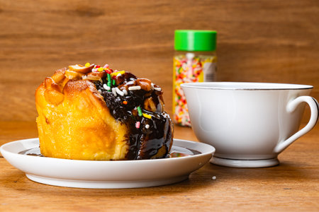 View of delicious butter bun garnished with nuts and rainbow sprinkles pouring with chocolate syrup in white ceramic dish and a white coffee cup on wooden table.の写真素材