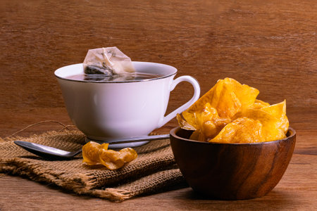 Sweet crispy potato chips in wooden bowl and a cup of tea with tea bag in white ceramic cup and metal spoon on sack cloth placed on wooden table.の写真素材