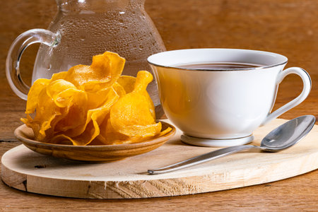 Closeup view of sweet crispy potato chips in wooden plate with a white ceramic cup of hot tea and a metal spoon on wooden cutting board and a transparent glass pitcher on wooden table.の写真素材