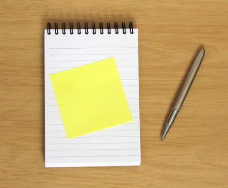 blank yellow postit stuck to spiral notebook with pen on wooden desk background. Add your own textの写真素材