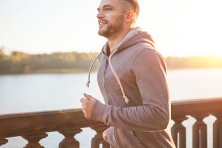 Young man running along embankment of river on sunset. Lens flare, warm tones.の写真素材