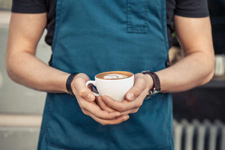 Barista in black t-shirt and blue apron holding  a cup of coffee with latte artの写真素材