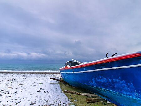Low angle view of snow covered beach and blue boat, under cloudy sky.の写真素材