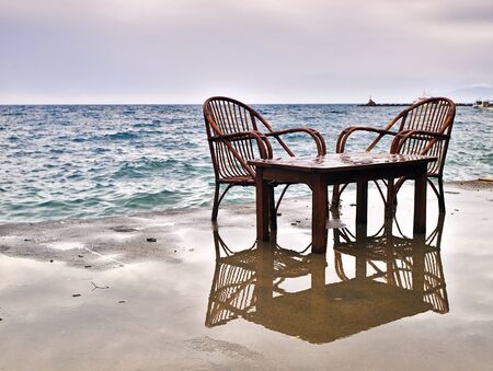 One table and two chairs left on seafront dock during stormy weather reflecting on wet surface. Of season concept.の写真素材