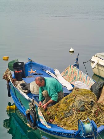 Kavala, Macedonia / Greece - May 14, 2019: Fisherman working on his fishing nets in traditional weathered blue wooden boat in the port of Kavala.のeditorial素材