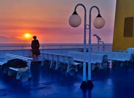 Young woman on a ship looking at the sunset, blue deck with white benches and yellow chimney, distant island mountains, sailing at Aegean sea, Greece.の写真素材