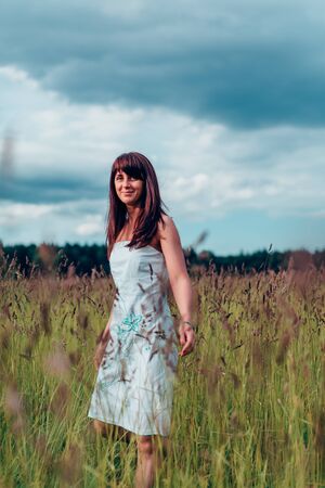 Beautiful happy brunette girl with her long hair in a white dress walks in the field.の写真素材