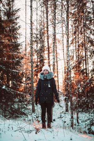 Pretty girl in a white hat and a jacket on a background of a winter forest. Photo girl in full growth.の写真素材