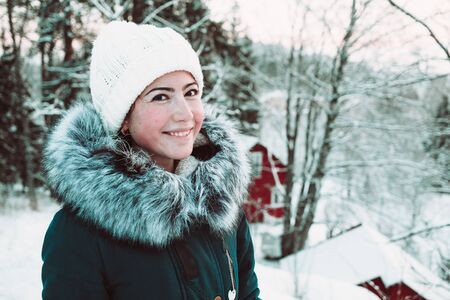 Beautiful young woman in a white cap and a jacket on a background of a winter forest. Close up photo.の写真素材