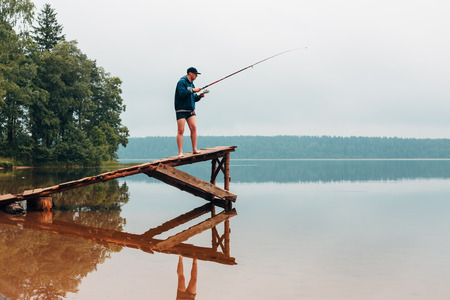 Man pulls a fishing rod standing on a wooden bridge. Weekend photo.の写真素材