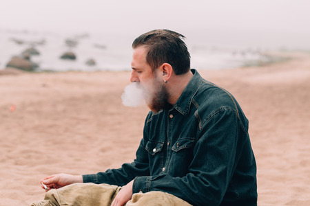Serious bearded man smoking a cigarette on the beach. bearded hipster on the beachの写真素材